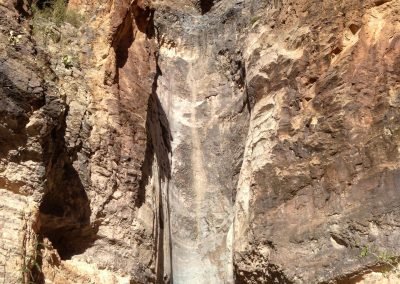 A towering rock formation with steep, textured cliffs, showing layers of sediment. A small waterfall cascades down the rock face, surrounded by dry vegetation and a clear blue sky overhead.