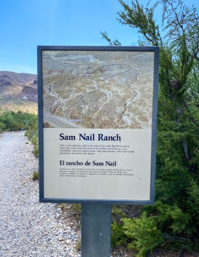 A sign at Sam Nail Ranch displaying information about the site. The sign is positioned on a gravel path, with a view of low desert vegetation and rocky terrain in the background. The text is visible in both English and Spanish.